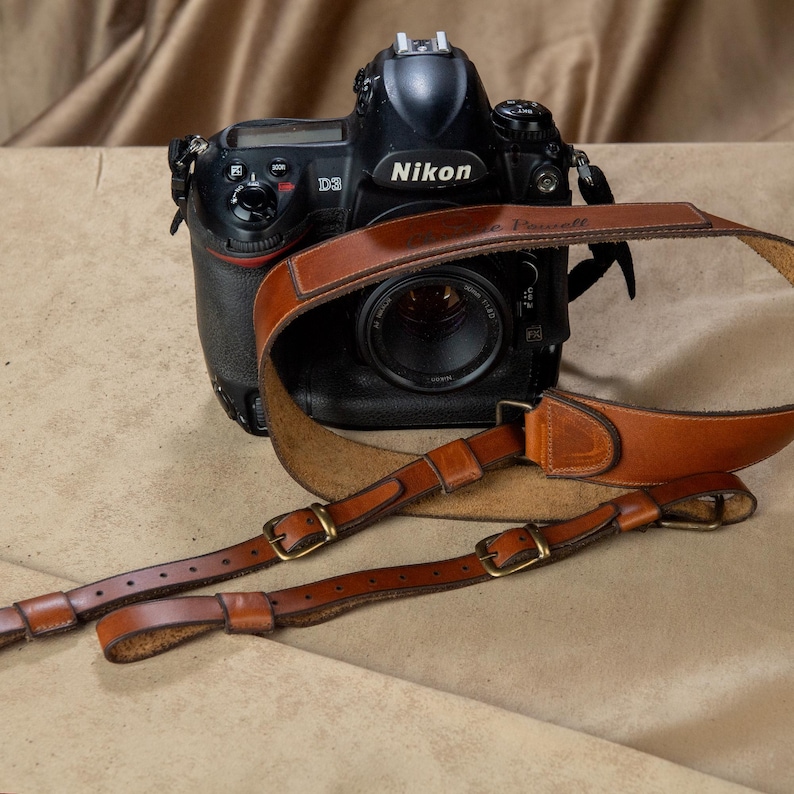 Op de afbeelding: Een zwarte Nikon D3 digitale SLR-camera met een bruinleren camerariem. De riem heeft messing gespen en ligt op een bruine ondergrond. De cameralens is zichtbaar.