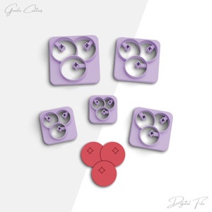 May include: Set of five purple plastic cookie cutters shaped like blueberries. The cutters are arranged in a pattern on a white background. There are three different sizes of cutters. The smallest cutter is in the center of the image. The largest cutter is on the left side of the image. The cutters are all in focus. There are three red plastic blueberries in the bottom center of the image.