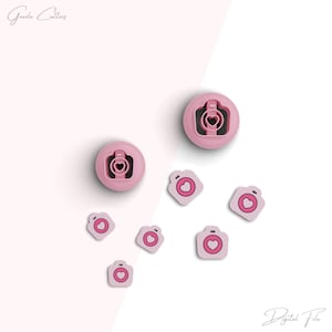May include: Pink cookie cutters in the shape of cameras with heart-shaped lenses. The image includes two larger circular cutters and several smaller, square-shaped cutters. The text "Graze Cutters" and "Digital File" are in the image.
