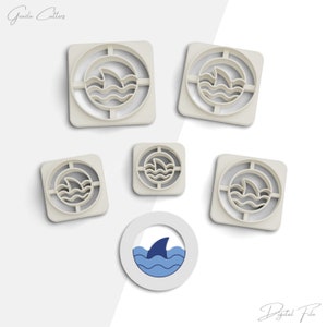 Puede incluir: Conjunto de cinco cortadores de galletas de diferentes tamaños, todos con una aleta de tiburón y olas dentro de un círculo. Los cortadores son blancos y de plástico. La imagen también muestra un diseño de aleta de tiburón y olas azul y blanco sobre un fondo blanco.