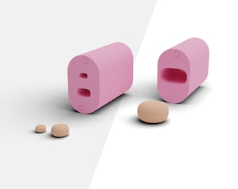 Puede incluir: Molde de plástico rosa con dos recortes redondos. El molde se muestra en dos vistas, una cerrada y otra abierta. La vista abierta muestra los recortes redondos. Hay tres pequeños objetos redondos de color beige junto al molde.