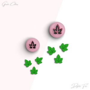 Puede incluir: Dos cortadores de galletas en forma de hoja de plástico rosa con contorno negro. Cinco cortadores de galletas en forma de hoja de plástico verde están esparcidos sobre una superficie blanca.