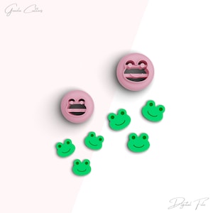 May include: A set of frog-shaped cookie cutters. There are two larger cutters with a pink frog face and two smaller cutters with a green frog face. The cutters are arranged on a white surface.