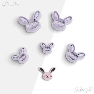 May include: Five purple plastic bunny cookie cutters of varying sizes. The smallest cutter has a pink face with two hearts. The largest cutter has a detailed bunny face with a nose and whiskers.