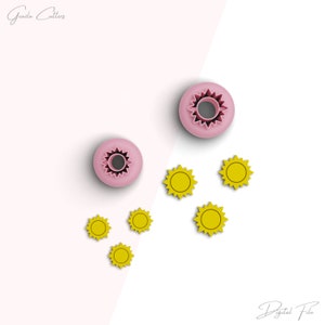 May include: Two pink cookie cutters shaped like a sun with a hole in the center. Five yellow cookie cutters shaped like a sun with a hole in the center.  The cookie cutters are on a white surface.