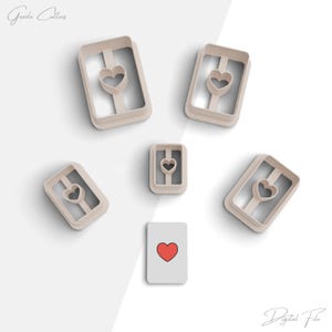 Puede incluir: Cinco cortadores de galletas en forma de carta de juego con un corazón en el centro. Los cortadores están hechos de plástico marrón claro. También hay una sola carta de juego con un corazón rojo en el centro.