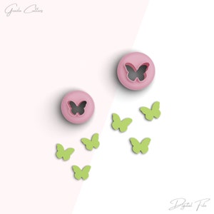 Könnte beinhalten: Zwei rosa Plastik-Ausstecher in Form eines Schmetterlings mit einem schwarzen Schmetterlingsdesign. Es gibt sechs grüne Plastik-Ausstecher in Form eines Schmetterlings, die um die Ausstecher herum verstreut sind.