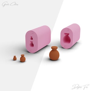 Op de afbeelding: Twee roze siliconen mallen voor het maken van kleine kleipotten. De mallen zijn aan één kant open en hebben een ontwerp van een kleine pot met een ronde buik en een smalle hals. Op de voorgrond zijn drie kleine kleipotten te zien, waarvan er één groter is dan de andere twee.