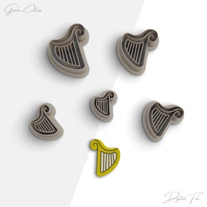 May include: Five cookie cutters in the shape of a harp. The cutters are made of a light brown plastic. The cutters are arranged in a scattered pattern on a white background. The largest cutter is in the upper left corner of the image. The smallest cutter is in the lower right corner of the image. The cutters are all the same design, but they are different sizes. The cutters are for making cookies in the shape of a harp.