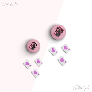 May include: Pink plastic cookie cutters in the shape of a camera with a heart inside. There are also five smaller white plastic cutters with a pink heart inside.