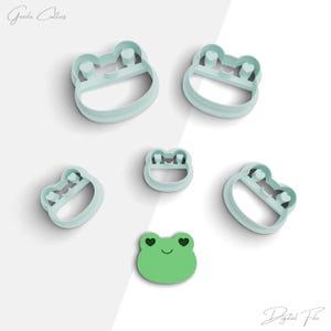 Può includere: Set di cinque stampini per biscotti a forma di rana di diverse dimensioni. Gli stampini sono di colore azzurro chiaro e presentano un viso di rana con occhi a forma di cuore. Lo stampino più piccolo è una rana verde piena con occhi a forma di cuore.