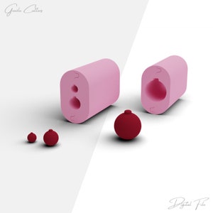 May include: A pink silicone mold for making ornaments. The mold is open and shows the shape of a round ornament with a small loop at the top. There are three red ornaments next to the mold.