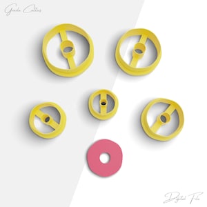 Pode incluir: Um conjunto de seis cortadores de donut de plástico amarelo, em vários tamanhos, e um cortador de donut rosa. Os cortadores são para fazer donuts, e o cortador é para decorar.