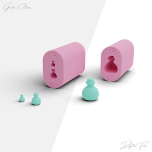 Puede incluir: Dos moldes de silicona rosa para hacer un pequeño objeto con forma de botella, redondo y tridimensional. Los moldes están abiertos y muestran el interior del molde. Hay tres pequeños objetos con forma de botella de color verde azulado en primer plano.