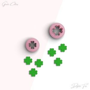 May include: Two pink plastic cookie cutters shaped like a four-leaf clover. Five green four-leaf clover shapes are scattered around the cutters.