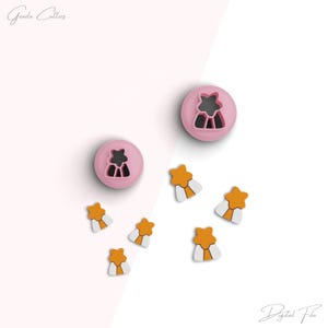 May include: Two pink cookie cutters, one shaped like a star and the other shaped like a tree, and six orange cookie cutters shaped like stars with white centers. The cookie cutters are on a white surface with a pink background.