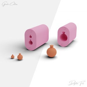 Op de afbeelding: Twee roze siliconen mallen voor het maken van kleine, ronde, keramische ornamenten. De mallen zijn open en laten de vorm van de ornamenten zien. Drie kleine, ronde, oranje ornamenten zijn te zien naast de mallen.