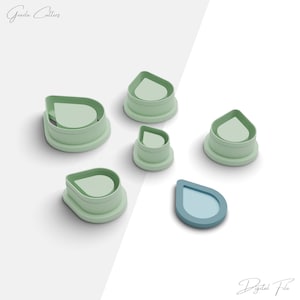 Puede incluir: Conjunto de cinco cortadores de galletas de plástico verde en forma de lágrima. Los cortadores están dispuestos en semicírculo sobre una superficie blanca. El cortador más pequeño está en el centro del semicírculo. El cortador más grande está en la parte superior del semicírculo. Todos los cortadores tienen la misma forma, pero son de diferentes tamaños. Hay un cortador de galletas en forma de lágrima azul en la esquina inferior derecha de la imagen.