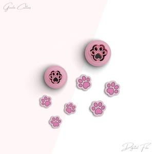 Pode incluir: Cortadores de biscoitos de plástico rosa em forma de pegadas de patas. Existem dois cortadores maiores e seis cortadores menores. Os cortadores maiores têm um desenho de pegada de pata dentro do cortador. Os cortadores menores são apenas pegadas de patas.