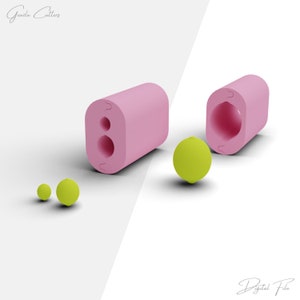 Könnte beinhalten: Rosa Silikonform zum Herstellen von runden und ovalen Bonbons oder Pralinen. Die Form ist offen und zeigt das Innere der Form. Neben der Form befinden sich drei grüne Bonbons.