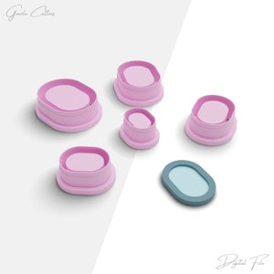 May include: Five pink oval-shaped cookie cutters and one blue oval-shaped cookie cutter. The cookie cutters are arranged on a white surface.