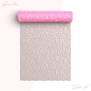 May include: A pink rolling pin with a textured surface of small circles. The rolling pin is unrolled, revealing a light grey surface with a repeating pattern of small circles.
