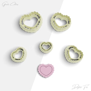 May include: Set of five heart-shaped cookie cutters in various sizes. The cutters are made of white plastic and have a scalloped edge. The smallest cutter is filled with a pink heart.