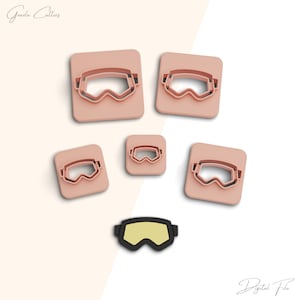 May include: Set of five pink plastic cookie cutters shaped like goggles. The cutters are in different sizes. There is also a black plastic goggle cookie cutter with a yellow lens.
