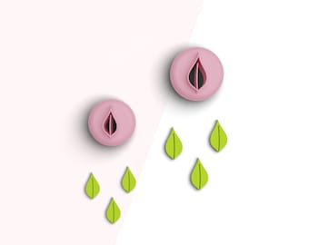 May include: Two pink cookie cutters shaped like a leaf with a small hole in the center. Five green leaf cookie cutters are scattered around the pink cutters.