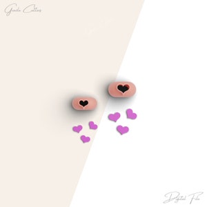May include: Two pink heart-shaped cookie cutters with a black heart cutout. There are six small purple hearts scattered around the cutters.