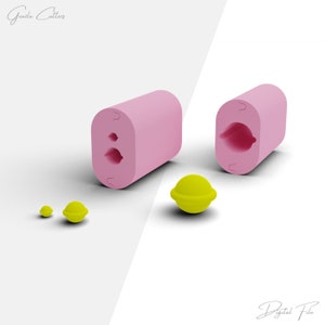 May include: Pink silicone mould with two cavities shaped like lemons. The mould is open and shows the inside of the cavities. There are three small yellow lemon-shaped sweets next to the mould.