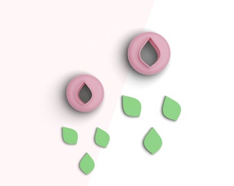May include: Two pink cookie cutters shaped like a teardrop with a smaller teardrop inside. Five green leaf-shaped cookie cutters are scattered around the pink cutters.