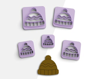 May include: Five purple plastic cookie cutters shaped like winter hats with a white pom-pom on top. One brown winter hat cookie cutter.