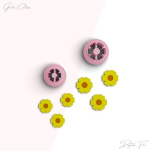 Pode incluir: Um conjunto de seis cortadores de biscoitos amarelos em forma de flor e dois cortadores de biscoitos rosa com um design de estrela de seis pontas. Os cortadores de biscoitos estão dispostos sobre uma superfície branca com um fundo rosa.