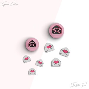 Op de afbeelding: Een set roze plastic koekjesvormen in de vorm van enveloppen met harten erin. De vormen zijn op een witte achtergrond met kleinere witte enveloppen met roze harten erin.