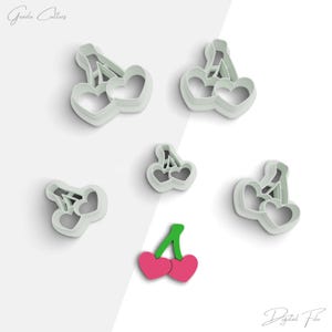 Puede incluir: Conjunto de seis cortadores de galletas con forma de cerezas. Los cortadores son blancos y tienen un tallo y dos cerezas en forma de corazón. También hay un cortador de cereza único con un tallo verde y dos cerezas rojas.