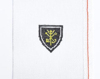 May include: A white needlepoint canvas with a black and yellow shield design. The shield features a yellow floral emblem on a black background, outlined in white. The canvas has a red border on the right side.