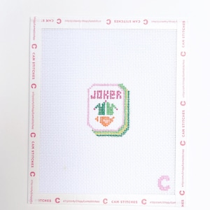 May include: A cross-stitch canvas featuring a colorful design of a playing card with the word "JOKER" in pink letters. The card has green, orange, and pink accents. The canvas is white with the shop name and logo printed around the edges.