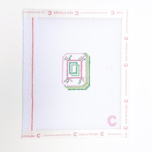 May include: A white needlepoint canvas with a geometric design in pink, green, and yellow. The canvas has a red vertical border on the left side and the text "CAM STITCHES" and "STITCHES" printed around the edges.
