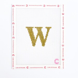 May include: A gold-colored, cross-stitch letter "W" on a white canvas. The canvas is bordered with pink text that reads "CAM STITCHES" and a website address. The letter is centered and stands out against the white background.
