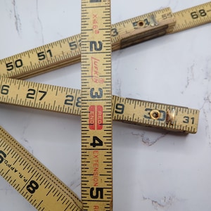 May include: A vintage wooden folding ruler with a red "JUFKIN" logo and the words "RED END" and "EXTENSION RULE" printed on it. The ruler is marked in inches and extends to 6 feet.