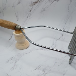 May include: A silver potato masher with a wooden handle. The handle is brown with a black stripe. The masher is resting on a small wooden stand.