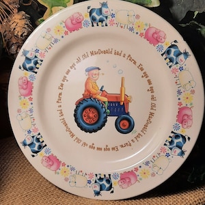 May include: A white ceramic plate with a colorful border of cows, pigs, sheep, and flowers. The center of the plate features a cartoon illustration of Old MacDonald driving a red tractor. The text "Old MacDonald had a farm. Eee eye eee eye oh!" is written around the tractor.