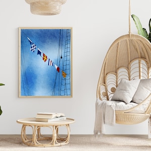 Puede incluir: Una pintura al agua enmarcada de un mástil de barco con banderas de colores contra un cielo azul. La pintura está colgada en una pared blanca sobre una silla colgante de mimbre con una manta blanca.