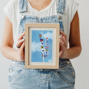 Op de afbeelding: Een ingelijste print van een nautisch vlagsignaal op een witte paal tegen een blauwe lucht met witte wolken. De vlaggen zijn rood, wit, blauw, geel en zwart.