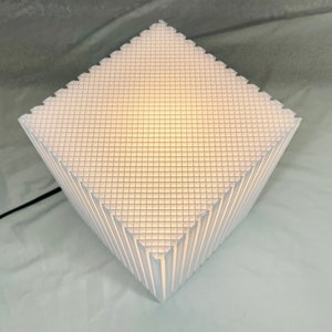 K&ouml;nnte beinhalten: Wei&szlig;e geometrische Tischlampe mit einem gitterartigen Muster. Die Lampe hat eine rautenf&ouml;rmige Form mit einer strukturierten Oberfl&auml;che und einem schwarzen Netzkabel. Die Lampe strahlt ein weiches, warmes Licht aus.