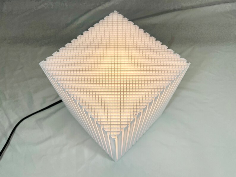 K&ouml;nnte beinhalten: Wei&szlig;e geometrische Tischlampe mit einem gitterartigen Muster. Die Lampe hat eine rautenf&ouml;rmige Form mit einer strukturierten Oberfl&auml;che und einem schwarzen Netzkabel. Die Lampe strahlt ein weiches, warmes Licht aus.