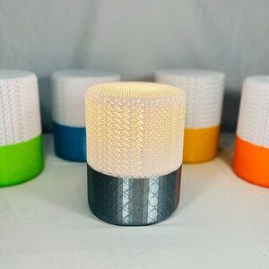 May include: A collection of cylindrical lamps with a white, textured top and a colored base. The lamps have a geometric pattern that emits a warm glow. The bases are in various colors, including green, blue, yellow, and orange.