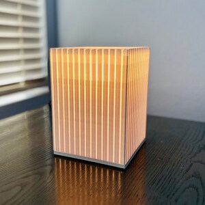 K&ouml;nnte beinhalten: Quadratische Lampe mit vertikalem Streifendesign, die ein warmes Licht ausstrahlt. Das Licht der Lampe scheint durch die vertikalen Lamellen. Sie steht auf einer dunklen Holzoberfl&auml;che. Abmessungen: ca. 20 cm x 20 cm x 20 cm.