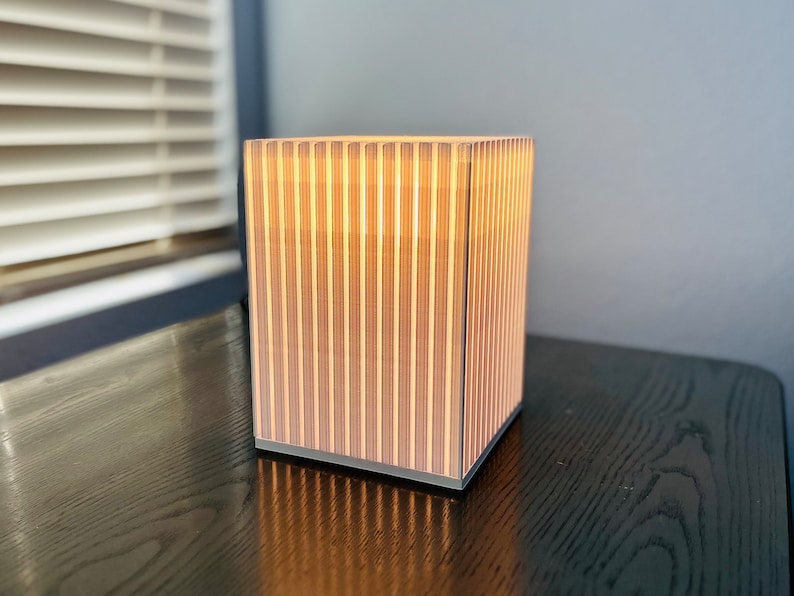 K&ouml;nnte beinhalten: Quadratische Lampe mit vertikalem Streifendesign, die ein warmes Licht ausstrahlt. Das Licht der Lampe scheint durch die vertikalen Lamellen. Sie steht auf einer dunklen Holzoberfl&auml;che. Abmessungen: ca. 20 cm x 20 cm x 20 cm.